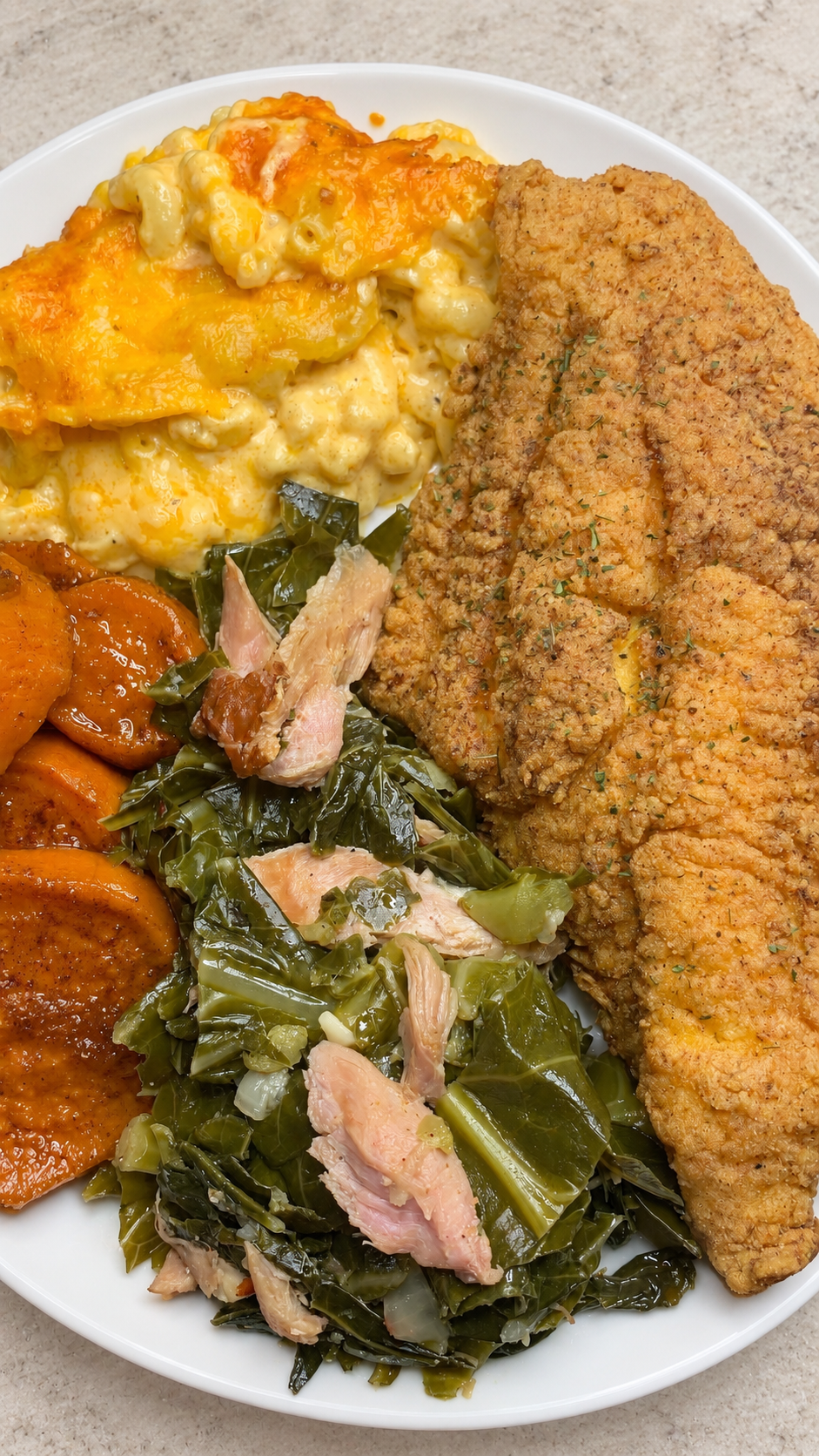 Fried catfish with baked macaroni and cheese, collard greens and candied yams on a white plate