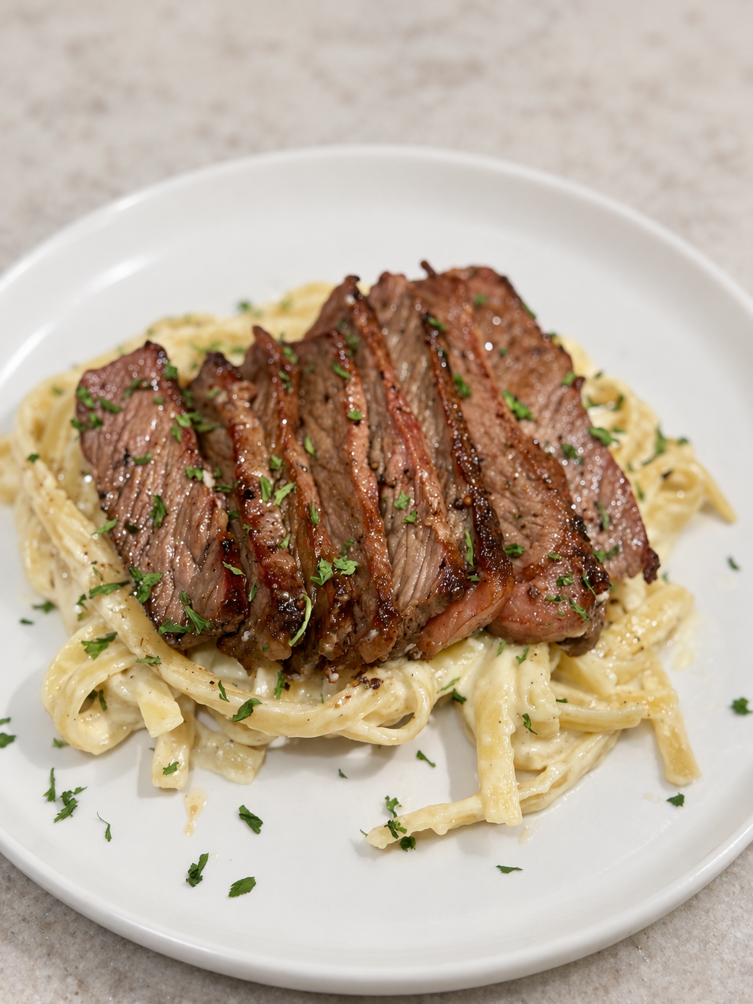 Sliced steak served over creamy Alfredo pasta on a white plate